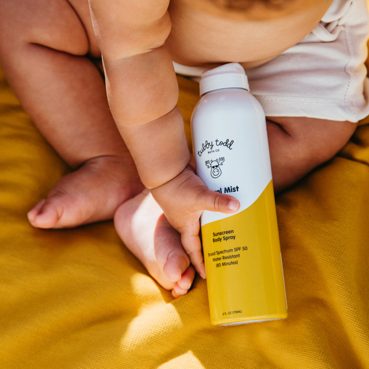 Close up of chubby baby holding Mineral Mist bottle on picnic blanket